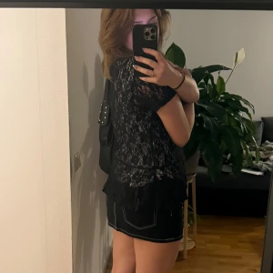 Svart spetsblus - Snygg svart spetsblus med korta ärmar och volangdetaljer. Perfekt för att ge en elegant touch till din outfit. Blusen har ett vackert blommönster och en rund halsringning. Passar bra till både kjol och byxor.