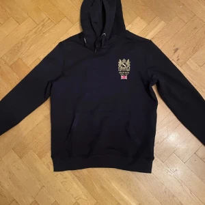 Marinblå hoodie från Morris - Snygg marinblå hoodie från Morris med broderad logga och Union Jack på bröstet. Perfekt för en stilren och bekväm look. Den har en klassisk känguruficka och justerbar huva med snören. Nypris 1300kr. Kontakta mig vid frågor 