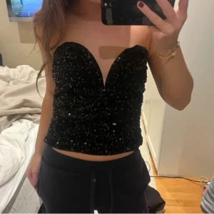 Svart paljettprydd tubtopp - Snygg svart tubtopp täckt med glittrande paljetter. Perfekt för en kväll ute eller festliga tillfällen. Toppen är axelbandslös och har en figurnära passform som framhäver silhuetten.