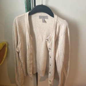 Beige kofta från H&M - Säljer en stilren beige kofta från H&M. Den har långa ärmar och knappar framtill, perfekt för lager på lager-stilen. Koftan är i ett mjukt material som gör den bekväm att bära. Passar utmärkt till både vardag och mer uppklädda tillfällen. Behöver bara strykas annars perfekt är den
