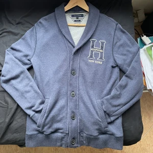 Blå kofta från Tommy Hilfiger - Säljer en snygg blå kofta från Tommy Hilfiger med stora knappar och ett broderat 'H' på bröstet. Koftan har långa ärmar och ribbade muddar vid ärmslut och nederkant. Perfekt för en stilren look!