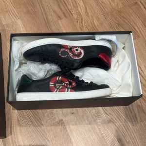 GUCCI ACE sneakers  - Snygga svarta sneakers från Gucci med ett iögonfallande ormmönster i rött och vitt på sidan. Skorna har en klassisk snörning och en vit sula som kontrasterar snyggt mot det svarta skinnet. Perfekta för den som vill ha en stilren men ändå unik look. Skick 9,5/10 bara lite smutsiga snören. Allt og tillkommer samt kvitto