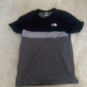 Svart och grå t-shirt från The North Face - Säljer en snygg svart och grå t-shirt från The North Face. T-shirten har korta ärmar och ett tryck med logotypen på bröstet. Perfekt för en avslappnad stil.