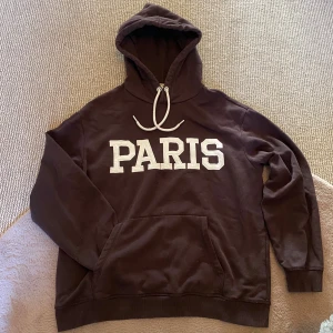 Hoodie - Säljer en brun hoodie från H&M med texten 'PARIS' i vitt på framsidan. Den har en klassisk känguruficka och justerbar huva med snören. Perfekt för en avslappnad stil.😻Bra att bara slänga på sig när du vill ha något mysigt❤️
