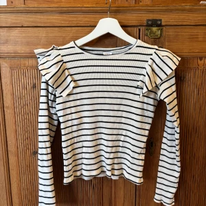 Randig långärmad tröja med volanger på axlarna  - Långärmad randig tröja i storlek 164, volanger på axlarna och ribbat tyg. Lite kortare i modellen så passar perfekt till ett par högre byxor. Sparsamt använd.