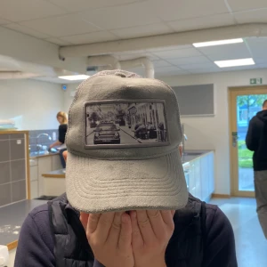 Grå keps från Emilio Pendenza - Säljer en snygg grå keps från Emilio Pendenza. Kepsen har en cool svartvit bild på framsidan och är tillverkad i ett mesh-material baktill för bra ventilation. Perfekt för en avslappnad stil!