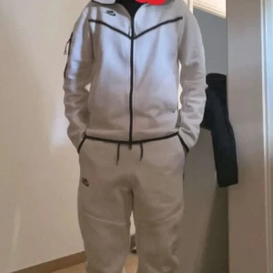 Vit Nike Tech Dress - Säljer en vit NIKE TECH Dress. Huvtröjan har dragkedjor framtill och är långärmad. Byxor med dragkedja på båda sidorna. Perfekt för en avslappnad stil. Dressen har svarta detaljer som ger en snygg kontrast. Skick 8/10