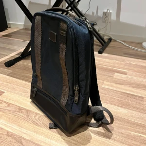 Blå ryggsäck med svarta detaljer - Säljer en snygg och praktisk blå ryggsäck med svarta detaljer. Den har flera dragkedjor och en stilren design som passar perfekt för både vardag och fest. Ryggsäcken är i bra skick och har justerbara remmar för bästa komfort.