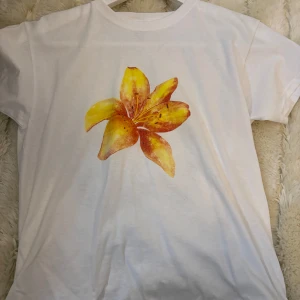 Vit t-shirt med blomtryck - Säljer en vit t-shirt med ett stort, färgglatt blomtryck i gult och orange på framsidan. Perfekt för sommardagar eller som en färgklick i garderoben. T-shirten är i bra skick och har korta ärmar.
