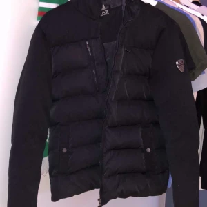 Svart pufferjacka från EA7 - Säljer en svart jacka frn EA7. Jackan har en quiltad design med dragkedja och knappar framtill. Perfekt för kalla dagar med sin varma och bekväma passform. 