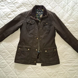 Barbour x Morris & co jacka vaxad bomull m William Morris foder strlk 34 - A brown Barbour jacket with a corduroy collar. The jacket has a zipper and snap buttons in the front, four front pockets with flaps and snap buttons, and a large inner pocket with a zipper. The jacket has a quilted lining and details in William Morri