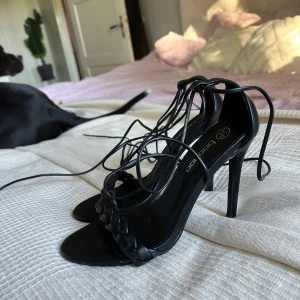 Svarta klackskor med snörning från Boohoo - Säljer ett par snygga svarta klackskor från Boohoo i storlek 39. De har en elegant flätad detalj framtill och långa snören som kan knytas runt vristen för en chic look. Perfekta för festliga tillfällen!