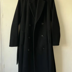 Svart kappa från Mint & Berry - Säljer en elegant svart kappa från Mint & Berry i storlek 38. Den är tillverkad av 50% ull och 50% polyester. Kappan har långa ärmar och en stilren design med knappar framtill och ett bälte i midjan. Perfekt för höst och vinter!