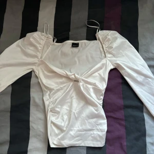 Vit topp från Gina Tricot - Säljer en superfin vit topp från Gina Tricot i storlek XXS men passar också till någon som har XS. Den har långa ärmar och en snygg knytdetalj framtill. Materialet är glansigt och känns lyxigt, perfekt för festliga tillfällen!