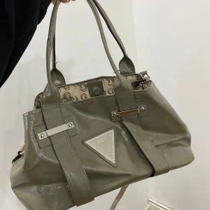 Grå handväska från Guess - Säljer en snygg grå handväska från Guess. Väskan har en glansig yta och är dekorerad med ett triangulärt Guess-emblem med små pärlor. Insidan är mönstrad med Guess-loggor och har en praktisk dragkedja. Perfekt för både vardag och fest!