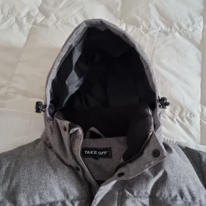 Grå pufferjacka från Take Off - Säljer en grå pufferjacka från Take Off. Den är perfekt för kalla dagar med sin tjocka vaddering och huva. Jackan har långa ärmar och stängs med både dragkedja och knappar. Den har också praktiska fickor framtill. Perfekt för vintern!