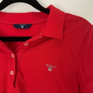 Röd pikétröja från GANT - Säljer en snygg röd pikétröja från GANT. Den har en klassisk krage och knappar framtill. Perfekt för en casual look eller en dag på golfbanan. Tröjan är i bra skick och redo för nya äventyr!