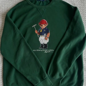 Grön sweatshirt med Polo Bear från Ralph Lauren - Säljer en grön sweatshirt från Ralph Lauren med den ikoniska Polo Bear på framsidan. Tröjan är i bra skick och har långa ärmar. Perfekt för en avslappnad stil eller som ett statement-plagg. Storleken är bekväm och passar de flesta.
