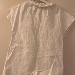 Vit blus från COS - Säljer en stilren vit blus från COS i storlek 36. Den har en hög krage med volangdetaljer och korta ärmar. Perfekt för både vardag och festliga tillfällen. Materialet är lätt och bekvämt, vilket gör den idealisk för vår och sommar.