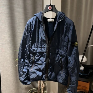 Mörkblå vindjacka från Stone Island - Säljer en snygg mörkblå vindjacka från Stone Island junio. Jackan har en dragkedja framtill och en huva för extra skydd mot väder och vind. Den är perfekt för höst och vår, den enda skadan är ett pytte litet hål i inner luv. Jackan är i använt skick 