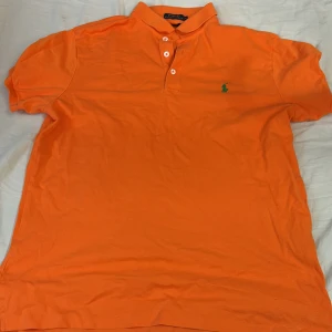 Orange pikétröja från Ralph Lauren - Säljer en snygg orange pikétröja från Ralph Lauren. Den har en klassisk krage med tre knappar och en liten grön broderad logga på bröstet. Tröjan är i ny skick men med ett litet håll i sidan.priset kan sänkas vid snabb affär 