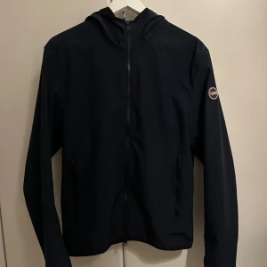 Svart vindjacka från Colmar - Säljer en stilren svart vindjacka från Colmar. Jackan har en dragkedja framtill och en praktisk huva. Perfekt för höst- och vårväder. Den är i bra skick och har en snygg logga på ärmen. Passar både till vardags och lite mer uppklädda tillfällen.