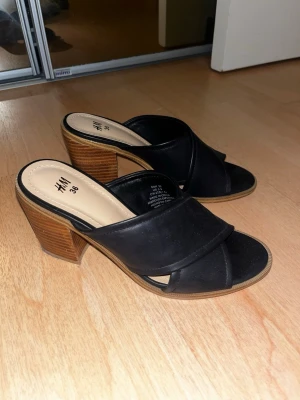 Svarta slip-in sandaler med klack H&M - Säljer ett par svarta slip-in sandaler från H&M med breda korslagda remmar och öppen tå. Klacken är chunky och träfärgad vilket ger en snygg kontrast till det svarta. Tillverkade i konstläder med bekväm innersula. Perfekta för en trendig look.