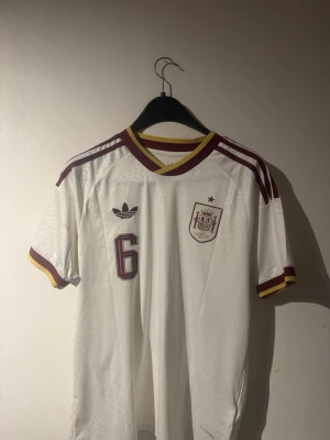 Vit Spanien borta fotbollströja Adidas - Vit fotbollströja från Adidas med Spaniens landslagsemblem och nummer 6 på bröstet. Baksidan har namnet Merino och nummer 6.