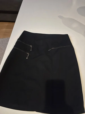 Svart kortkjol med dragkedjor - Stilren svart kortkjol med två sneda dragkedjor framtill som ger en cool detalj. Kjolen har en enkel, rak passform och är perfekt att matcha med olika toppar. Materialet ser ut att vara polyester eller liknande syntet.