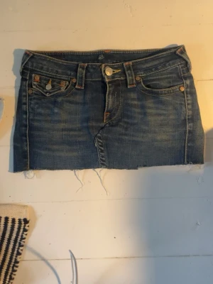 True religon jeanskjol - Säljer en kort true religon jeanskjol i blå denim med avklippt kant eftersom jag ville ha den kortare. Kjolen har klassiska fem fickor, synliga sömmar och knappgylf. Baksidan har de klassiska truey fickorna bak. Mammas gamla kjol, men fortfarande i gott skick!💗