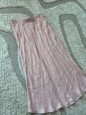 Ljusrosa satinkjol från H&M - Säljer en lång, ljusrosa satinkjol från H&M. Kjolen har en mjuk glansig yta och en rak, flowy passform som ger en snygg siluett. Perfekt att styla med en enkel topp eller stickad tröja. Materialet känns svalt och följsamt.