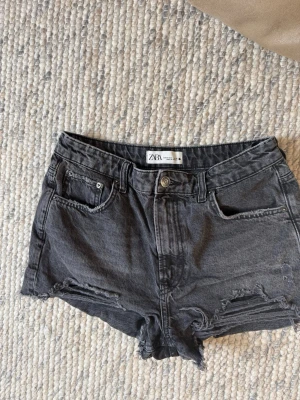 Svarta  jeansshorts Zara - Säljer ett par svarta högmidjade jeansshorts från Zara. Shortsen har råa kanter, slitningar framtill och klassisk femficksdesign. Perfekta till sommaren för en avslappnad och trendig look.