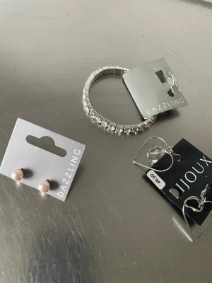 Smyckeset med örhängen och armband - Säljer ett set med smycken som inkluderar ett silvrigt armband med fyrkantiga stenar, ett par rosa pärlörhängen och två par silvriga örhängen. Märkena Dazzling och Bijoux. Perfekt för att mixa och matcha till olika looks.