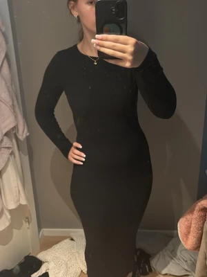 Svart långärmad långklänning - En stilren svart, ribbad, långklänning med lång ärm och rund halsringning. Klänningen har en tight passform som framhäver figuren och är perfekt för dig som gillar en enkel men elegant look. Helt oanvänd 