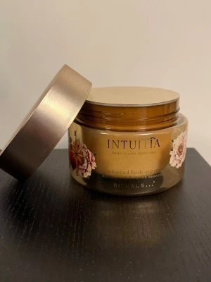 Rituals Intuitia whipped body cream - Lyxig whipped body cream från Rituals i serien Intuitia med doft av sandelträ och vanilj. Burken är guldbrun med blommiga detaljer och har ett elegant lock i metallic. Perfekt för dig som vill ha en återfuktande och väldoftande kroppskräm. Ny och oöppnad!