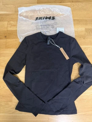 Svart långärmad topp från Skims - En svart långärmad topp från Skims i mjuk bomullsmix. Enkel och stilren design med rund halsringning och figurnära passform. Perfekt att styla med jeans eller kjol för en clean look.