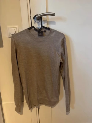 Beige stickad tröja från Oscar Jacobsson  - Säljer en stilren beige stickad tröja från Oscar Jacobsson med rund halsringning och långa ärmar. Tröjan är tillverkad i en mjuk ullmix och har ribbade muddar vid ärmslut och nederkant. Perfekt för lager på lager under kyliga dagar.