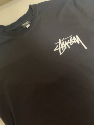 Svart Stüssy t-shirt med logga - Säljer en svart t-shirt från Stüssy med den klassiska vita loggan tryckt på bröstet. T-shirten har rund halsringning och korta ärmar. Perfekt för dig som gillar streetwear och vill ha en enkel men snygg look.