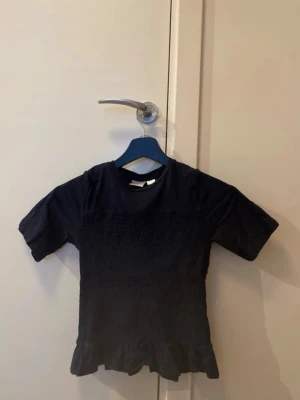 Svart topp med volangkant - Säljer en svart topp med korta ärmar och smockad midja som ger en tight passform. Toppen har en söt volangkant nedtill och är enkel men ändå stylish. Perfekt att matcha med jeans eller kjol för en trendig look.