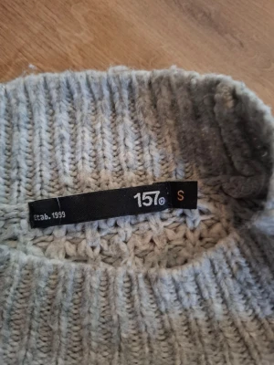 Ljusgrå stickad tröja från 157 - Mysig ljusgrå stickad tröja från 157 med grovstickad struktur och ribbad hals, ärmslut och nederkant. Perfekt för lager på lager och chill dagar. Tröjan har en avslappnad passform och är tillverkad i mjukt material.