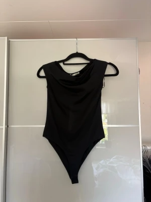 Svart ärmlös body med draperad hals - Snygg svart body med draperad halsringning och ärmlös design från Zara. Tillverkad i ett stretchigt, mjukt material. Perfekt att styla till både jeans och kjol för en trendig look.