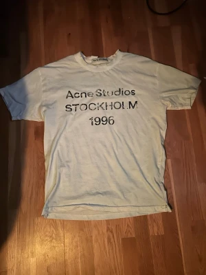 Vit Acne Studios t-shirt Stockholm - Vit t-shirt från Acne Studios med tryck 'Acne Studios STOCKHOLM 1996' på framsidan. Klassisk passform med rund hals och korta ärmar. Perfekt för dig som gillar minimalistisk och stilren design. Den e lite ljus blå