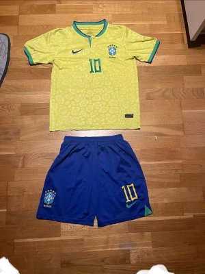 Brasilien matchställ fotboll gul/blå Neymar - Säljer ett Brasilien matchställ med gul tröja och blå shorts. Tröjan har gröna detaljer och nummer 10 på bröstet, samt ett subtilt mönster. Shortsen är blå med nummer 10, Nike-logga och Brasilien-emblem. Perfekt för fotbollsträning eller match.