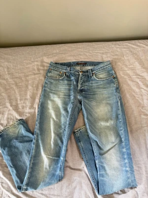 Nudie jeans Grim Tim - Säljer ett par ljusblå jeans från Nudie Jeans med raka ben och klassisk femficksmodell. Jeansen har slitningar och snygg tvätt, samt broderade bakfickor och läderpatch i midjan. Perfekta för en avslappnad och trendig stil.