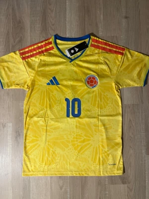 Colombia matchtröja Rodriguez #10 - Gul Colombia matchtröja med blå detaljer och röda ränder på axlarna. Nummer 10 och namnet 'James' på ryggen. Broderad landslagssköld på bröstet. Tillverkad i lätt och ventilerande material, perfekt för fotboll. HELT NY!
