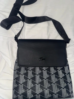 Svart axelväska  - Snygg svart axelväska med justerbar axelrem. Väskan har ett lock i svart konstläder och mönstrad framsida med vita geometriska linjer och Lacoste-loggor. Perfekt för dig som vill ha en stilren och modern look.