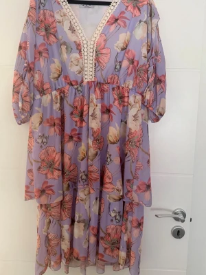 💜🌸 Ljuslila klänning med blommönster 🌸💜 - 💜🌸 Ljuslila klänning med blommönster 🌸💜  ✨ Köpt under en livesändning från Mili Made in Italy ✨ Tillverkad i ett mjukt och luftigt material med fina volanger 🌿 Dekolletaget är dekorerat med vit spets (gipyr) 🤍 Effektfulla ärmar med två öppningar som är sammanbundna med dekorativa snören 🎀 samt resårdetaljer ✨  🌸 Mycket fint skick — använd max 3 gånger 🤍  📏 Mått: • bredd: 130 cm (kan töjas till ca 140 cm) • längd: 103 cm  📌 Storlek: XL/XXL