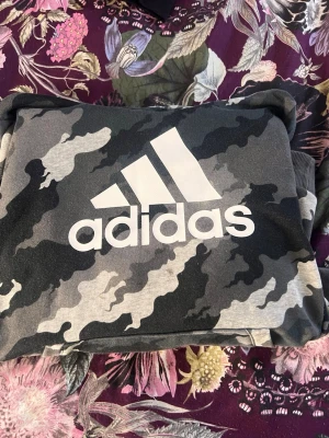 Camo sweatshirt från Adidas - Säljer en snygg sweatshirt från Adidas med grått och svart camouflagemönster och stor vit logga på bröstet. Perfekt för träning eller chill. Mjuk bomullskänsla och ribbade muddar vid ärmslut och nederkant.