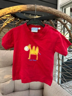 Röd t-shirt med kamel från Snuggles - Söt röd t-shirt från Snuggles med korta ärmar och tryck av en gul kamel på framsidan. T-shirten har praktiska knappar på axeln för enkel påklädning. Perfekt för små barn som gillar färgglada motiv.
