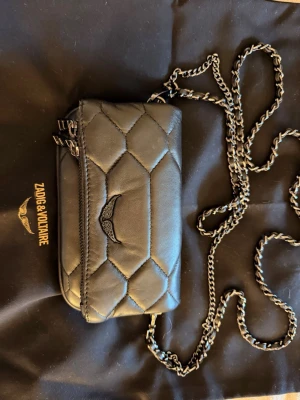 Svart quiltad axelväska Zadig & Voltaire - Snygg svart quiltad axelväska från Zadig & Voltaire med kedjerem och dubbla dragkedjor. Väskan har ett coolt vingemblem framtill och är tillverkad i mjukt läder. Perfekt storlek för det viktigaste och ger en trendig touch till din outfit.Allt medkommer, även digitalt kvitto 🌸🌸